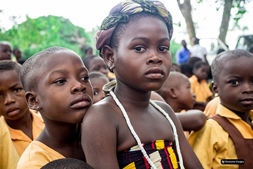 Young girl wearing head scarf and white necklace in a group of children from Ghana staring directly at camera. 