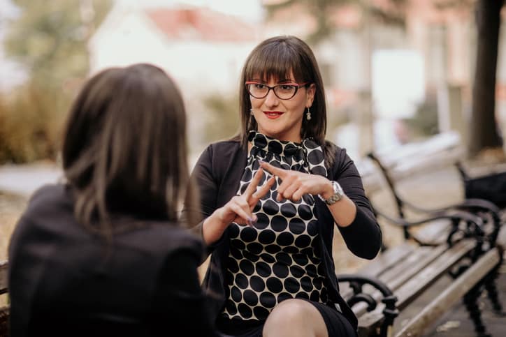woman in black dress and glasses performing sign language to another woman on a bench