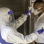 Nurse in PPE suit uses infrared thermometer measuring temperature with African male worker scanning for Coronavirus or Covid-19 symptom at office elevator International medical healthcare system