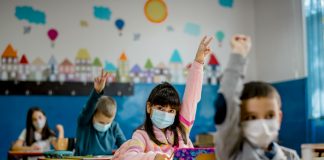 Kids in class with masks, raising their hands.