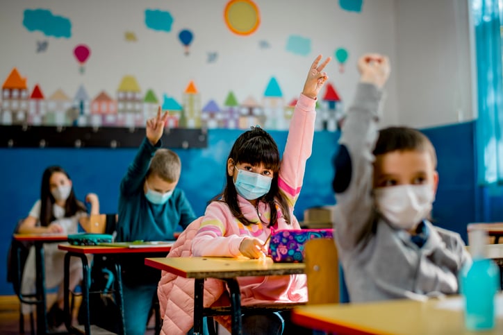 Kids in class with masks, raising their hands.