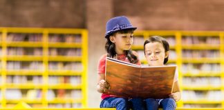 Two young kids sitting on a bright yellow platform reading a big storybook together