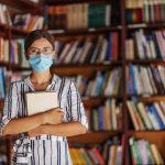 Portrait of young female librarian standing in library with face mask on holding a book.
