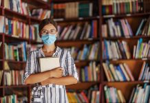 Portrait of young female librarian standing in library with face mask on holding a book.