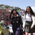 girl walking in school uniform with a friend who talks to the mobile with the town of colorful houses behind. Guanajuato, Mexico,