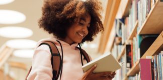 Smiling girl holding a book in library while standing at bookshelf
